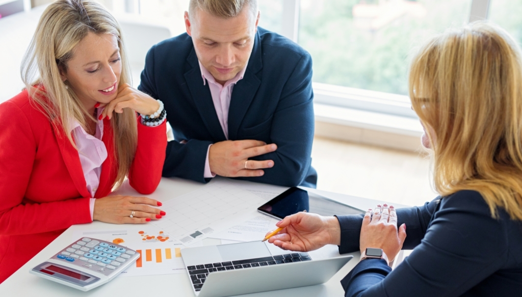 husband and wife discussing financial plans with an advisor. They are sitting around a table looking at a computer and documentation.