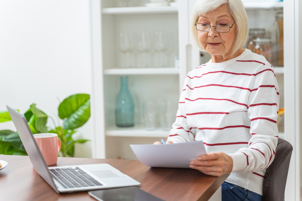 Elderly women sitting by a computer filling out paper work in her kitchen