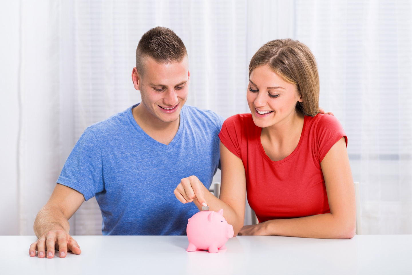 Couple sitting at a white table with a white background putting a coin into a pink piggy bank
