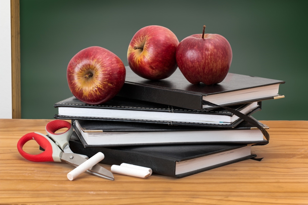 pile of black school books, apples, and scissors sitting on a deskg