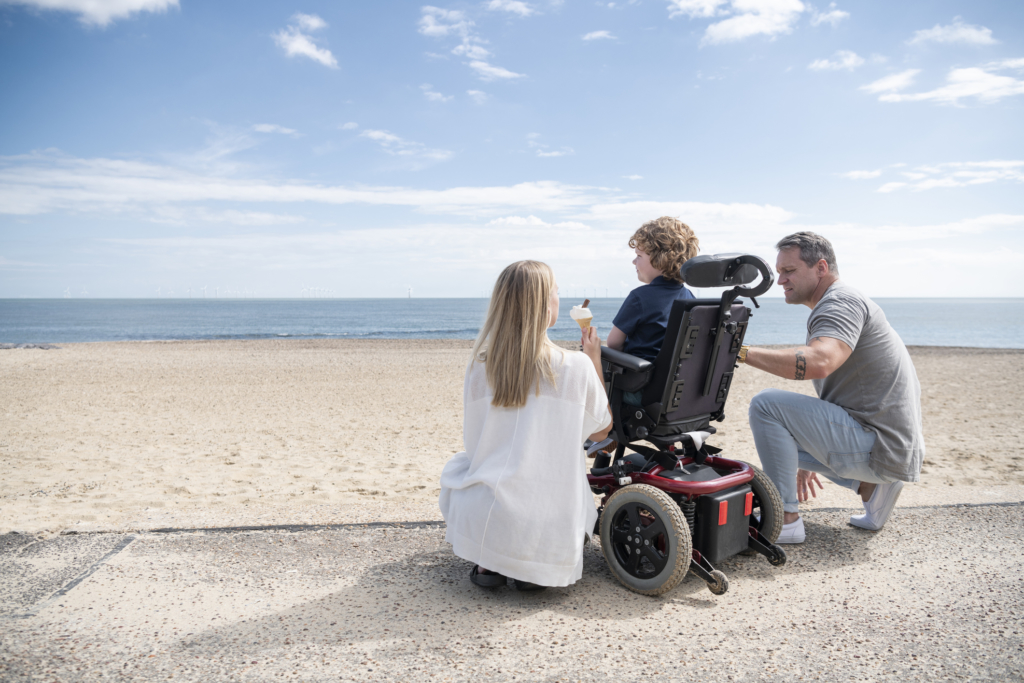 Mid adult mother and mature father kneeling by 6 year old child with muscular dystrophy by the beach, enjoying an ice cream while looking at the view. Clacton on Sea.