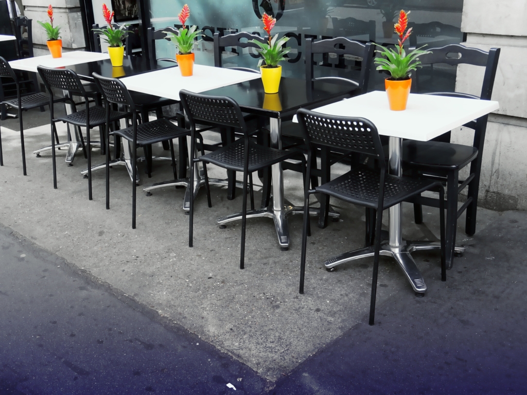Black chairs and black & white tables in a row on a patio in front of a restaurant. Potted flowers on each table..