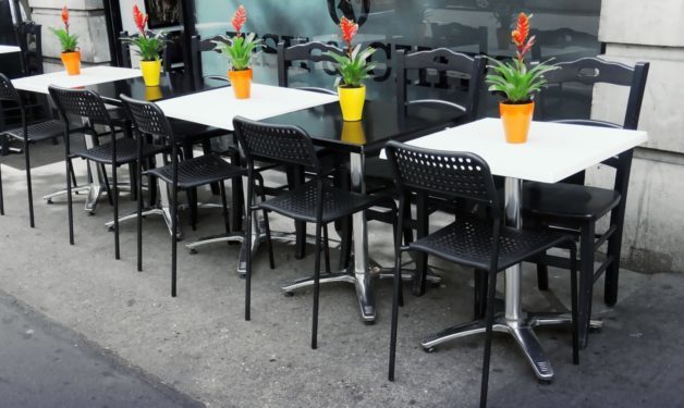 Black chairs and black & white tables in a row on a patio in front of a restaurant. Potted flowers on each table..