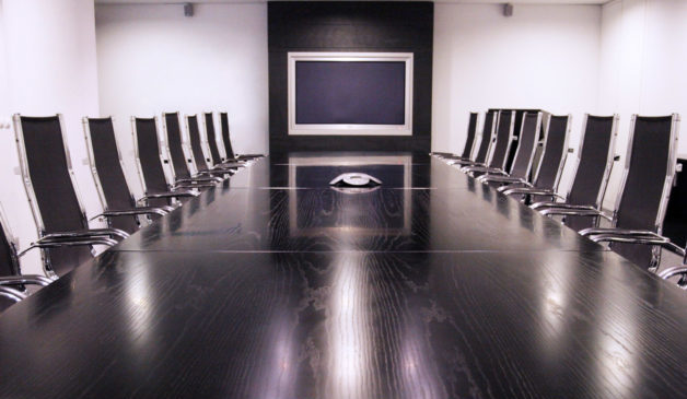 wooden boardroom table with black chairs around it. TV mounted on wall.