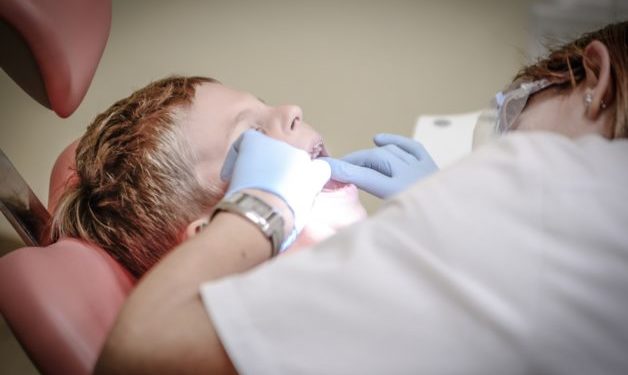 Child sitting at a red dentist chair. Dentist examining childs mouth.