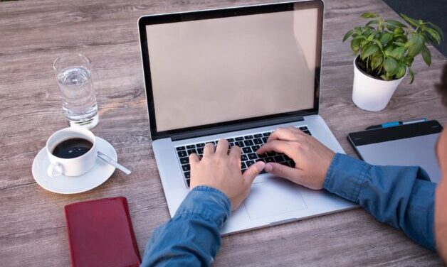 Close up of persons hands typing on a laptop. Person is wearing a blue shirt. The laptop is sitting on a wooden table with a coffee and plant beside it.