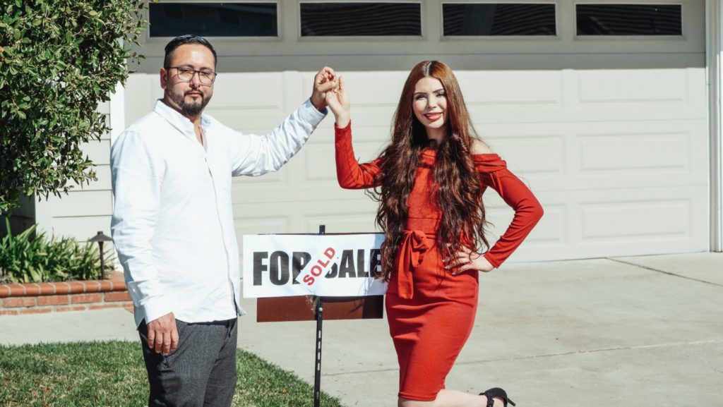 Photo of couple holding hands, standing in front of a sold sign and a house.