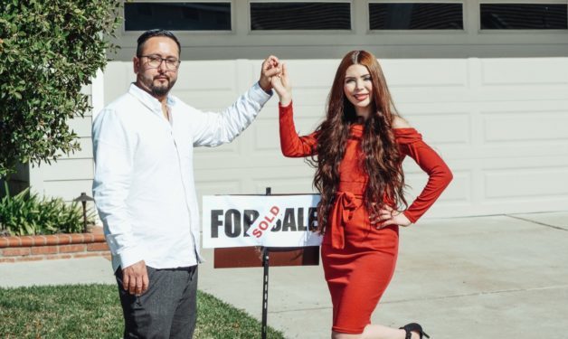 Photo of couple holding hands, standing in front of a sold sign and a house.
