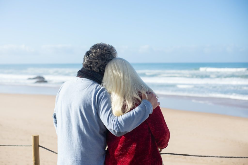 Older couple hugging, looking towards a beach. You can see the sand and the water.