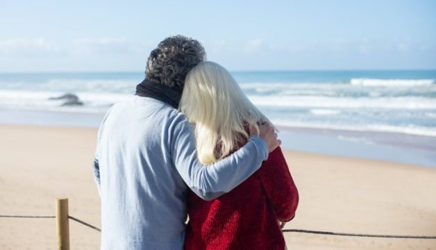 Older couple hugging, looking towards a beach. You can see the sand and the water.