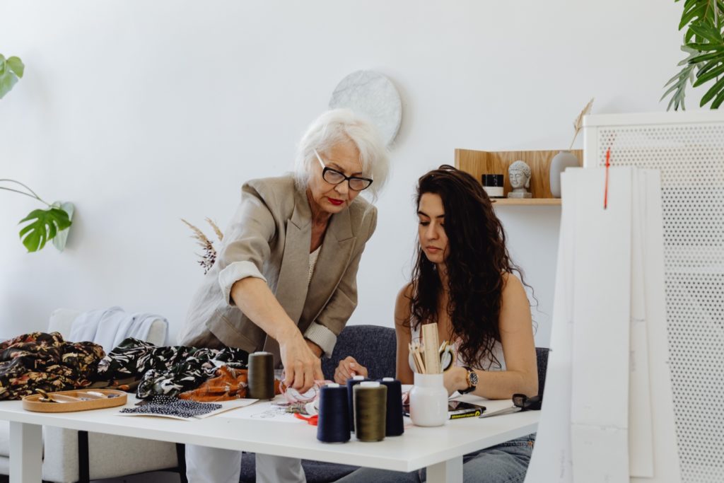 Older women dressed in a beige blazer, showing a younger female how to sew. Younger female is sitting at white desk with sewing equipment.