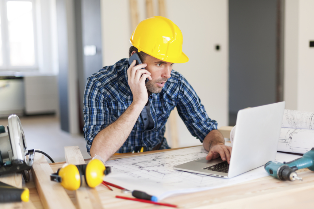 construction worker looking at laptop and sitting at his desk with his phone