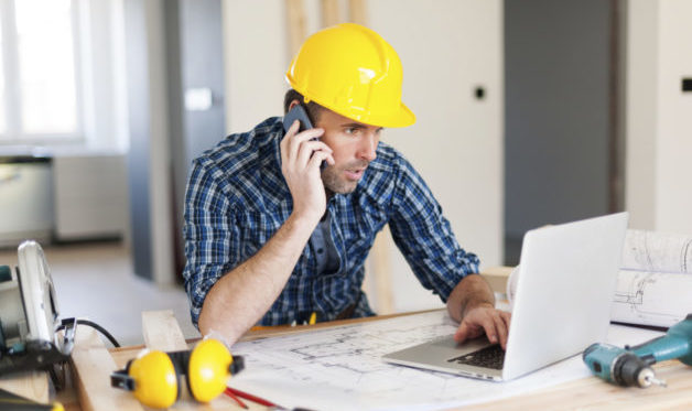 construction worker looking at laptop and sitting at his desk with his phone