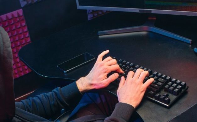 photo of computer with person typing. Person is wearing black hoodie over head, only hands visible.