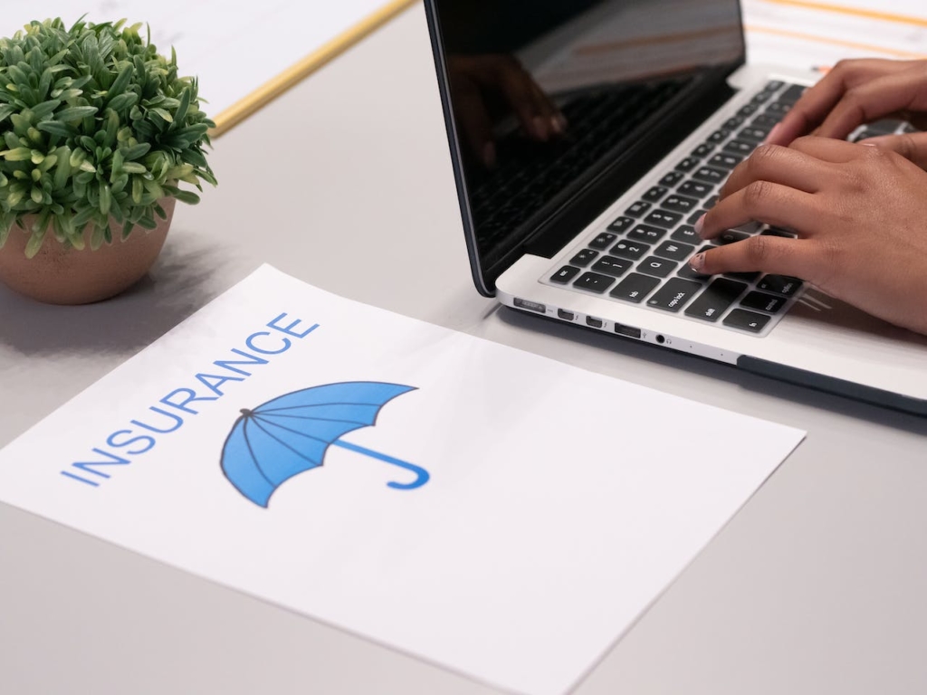 Photo of a desk with a piece of paper on it that says insurance. A blue umbrella underneath and a laptop to the side.