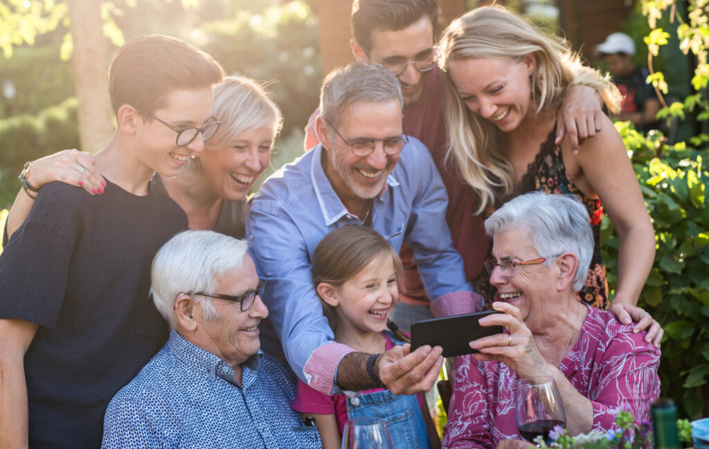 Summertime, a family with three generations having fun around a table in the garden sharing a meal. The whole guest having fun watching a video on a phone