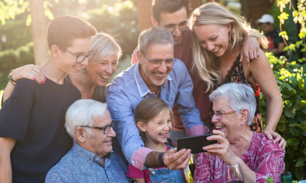 Summertime, a family with three generations having fun around a table in the garden sharing a meal. The whole guest having fun watching a video on a phone