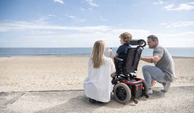 Mid adult mother and mature father kneeling by 6 year old child with muscular dystrophy by the beach, enjoying an ice cream while looking at the view. Clacton on Sea.