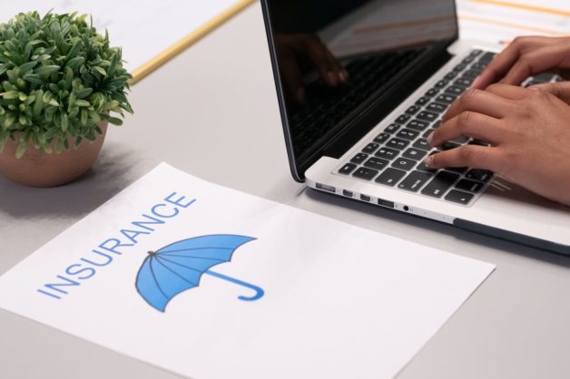 Photo of a desk with a piece of paper on it that says insurance. A blue umbrella underneath and a laptop to the side.