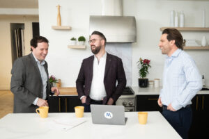 Ryan Bouskill, James Buckley, and Scott McGillivray laughing casually around a kitchen island with a laptop on the counter.