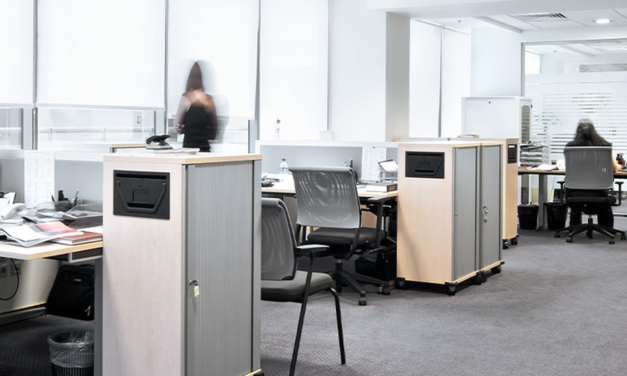 Office with multiple desks. People blurred in background. Office is white and gray.