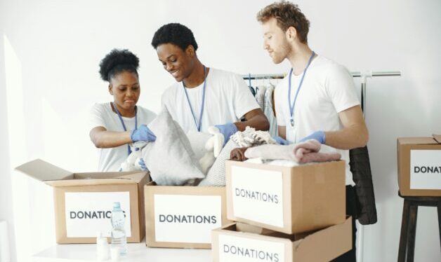 Picture of three people organizing a bunch of boxes for donations. The boxes are placed on a table, the background is white.