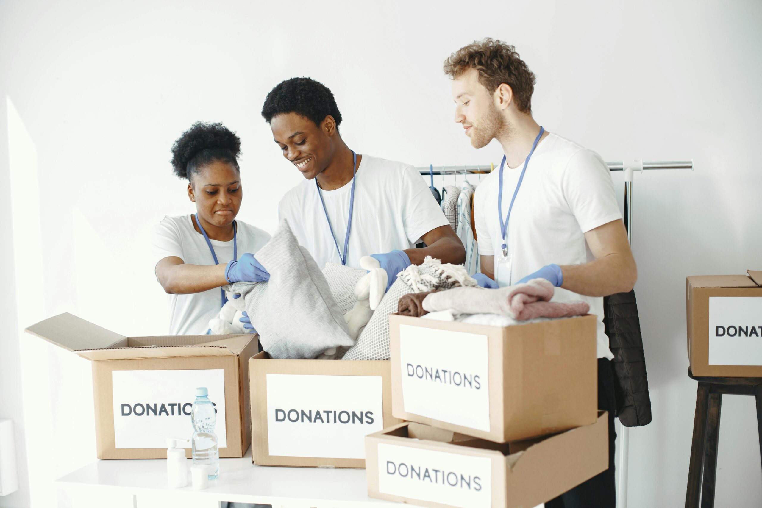 Picture of three people organizing a bunch of boxes for donations. The boxes are placed on a table, the background is white.