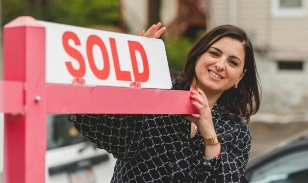 Realtor standing in front of a red sold sign. The realtor is female and wearing a blue sihirt.