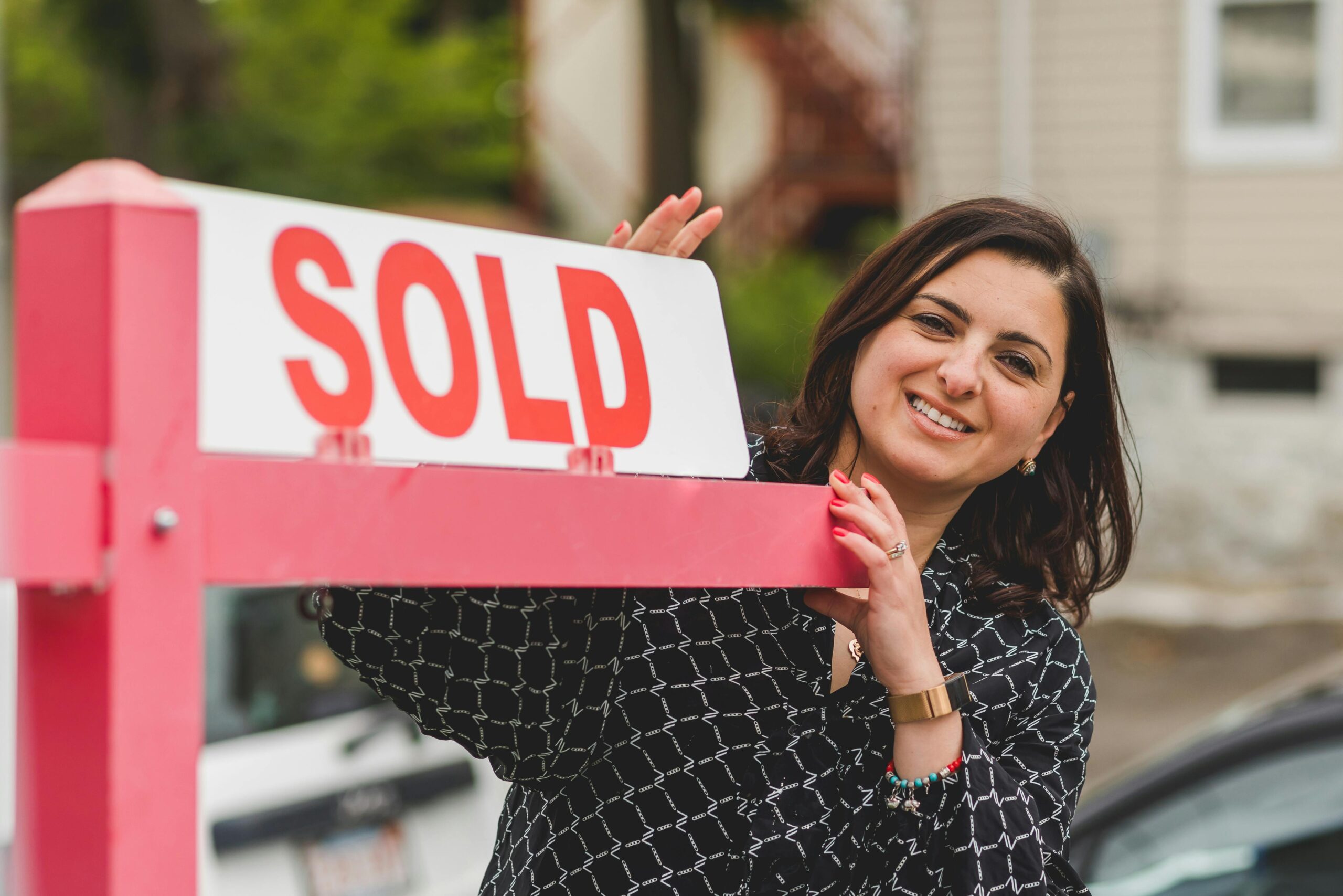 Realtor standing in front of a red sold sign. The realtor is female and wearing a blue sihirt.