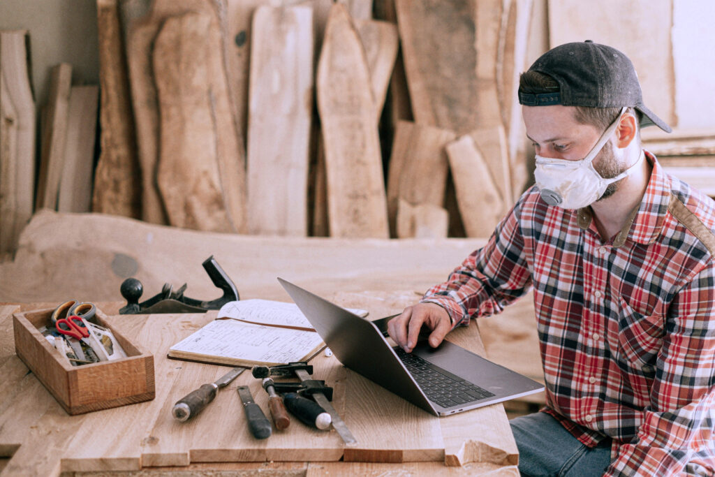 Photo of construction man in red plaid shirt, sitting at a desk, working on a laptop. There are construction tools sitting on the table.