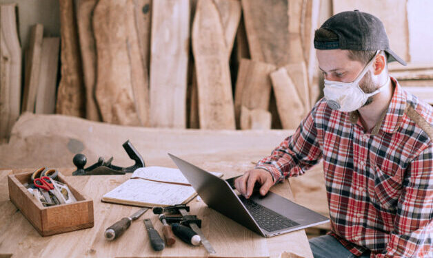 Photo of construction man in red plaid shirt, sitting at a desk, working on a laptop. There are construction tools sitting on the table.