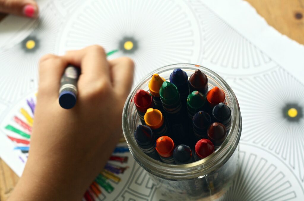 Close up of child's hand holding a blue crayon. Child is coloring a picture and has a container of crayons beside them.