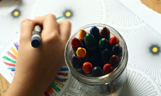 Close up of child's hand holding a blue crayon. Child is coloring a picture and has a container of crayons beside them.