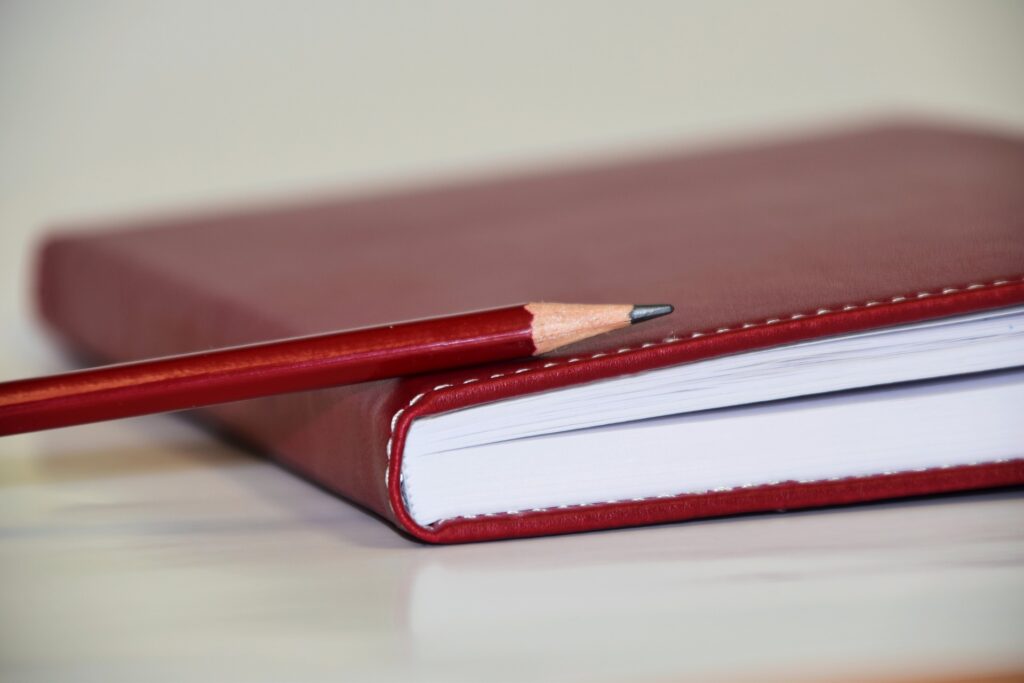 Picture of red notebook sitting on a white desk with a red pencil on top.