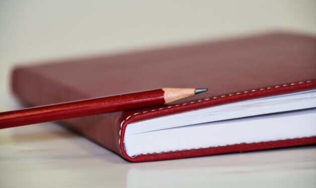 Picture of red notebook sitting on a white desk with a red pencil on top.