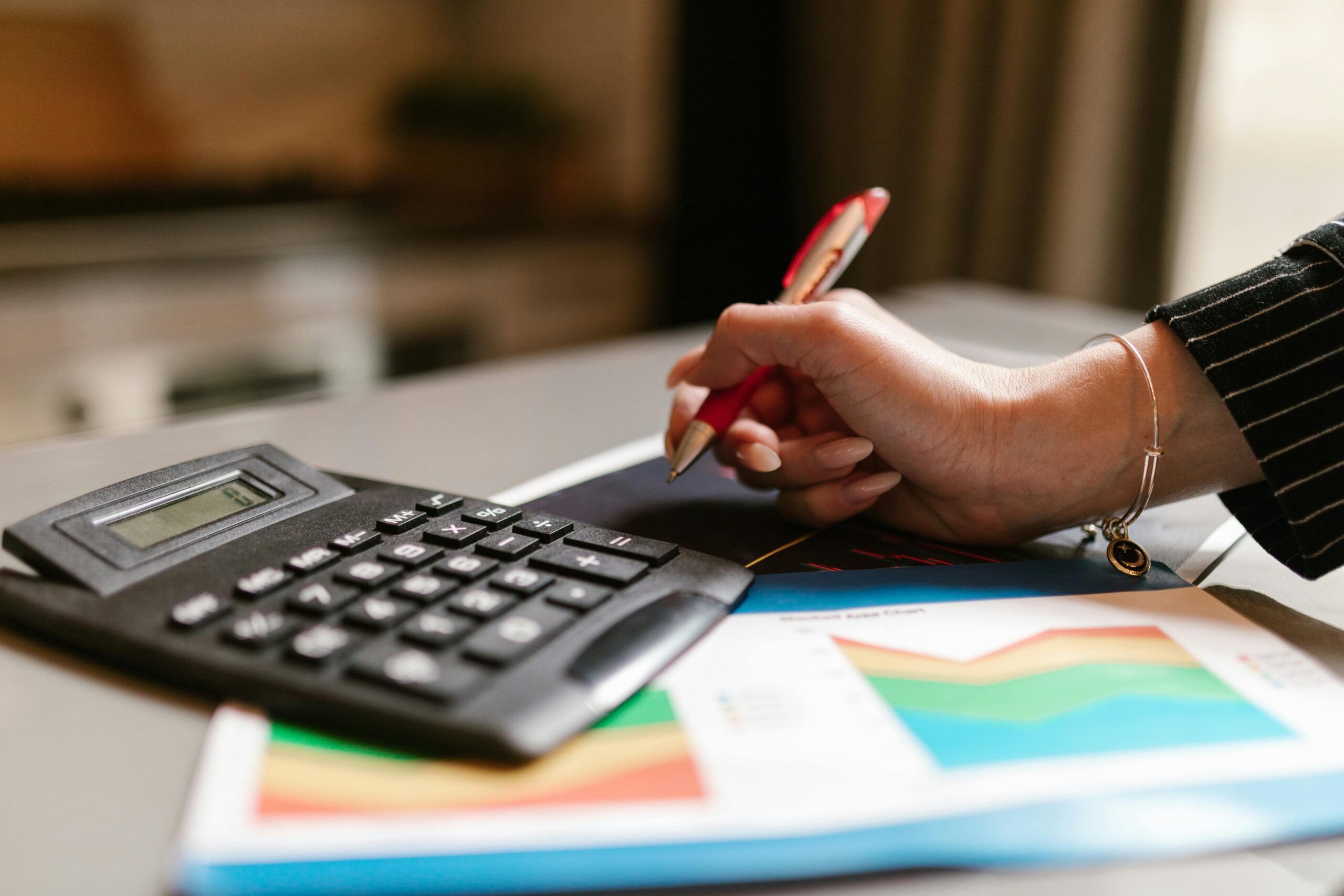 Close up of a desk with a calculator and paper work on it. Women's hand holding a red pen.