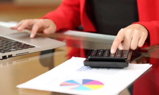 Close-up of women in red jacket and black shirt, using a calculator and laptop and sitting at a desk. There are some financial paper work on the desk.