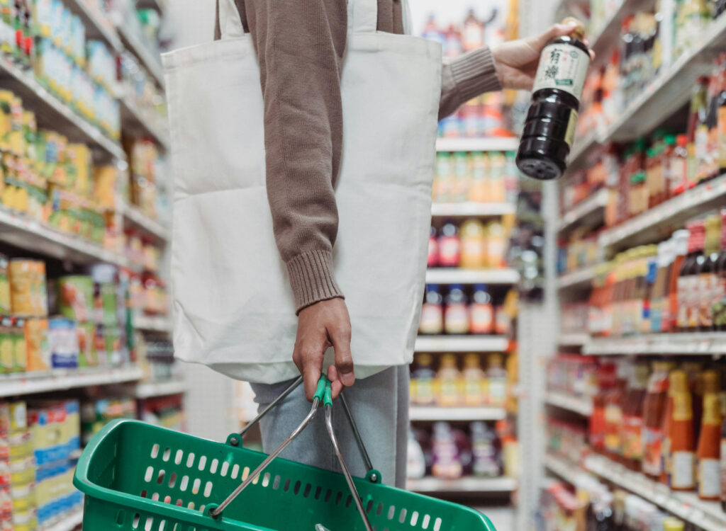 Close up of person grocery shopping, holding a green shopping basket and grabbing a bottle of the shelf. Only arm and leg of person is visible.