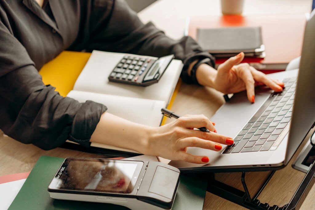 Closeup of women sitting at desk typing on a laptop. She is wearing a black shirt with the sleeves rolled up and has red nail polish. There is a calculator and notepad on the desk.