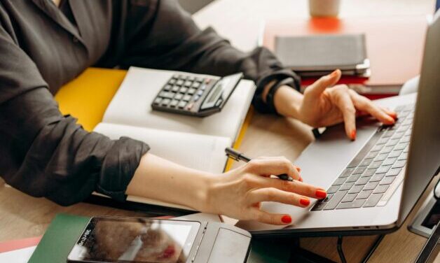 Closeup of women sitting at desk typing on a laptop. She is wearing a black shirt with the sleeves rolled up and has red nail polish. There is a calculator and notepad on the desk.