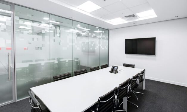 Empty boardroom. White table, black chairs, and a tv on the wall. One wall of the boardroom is made of glass.