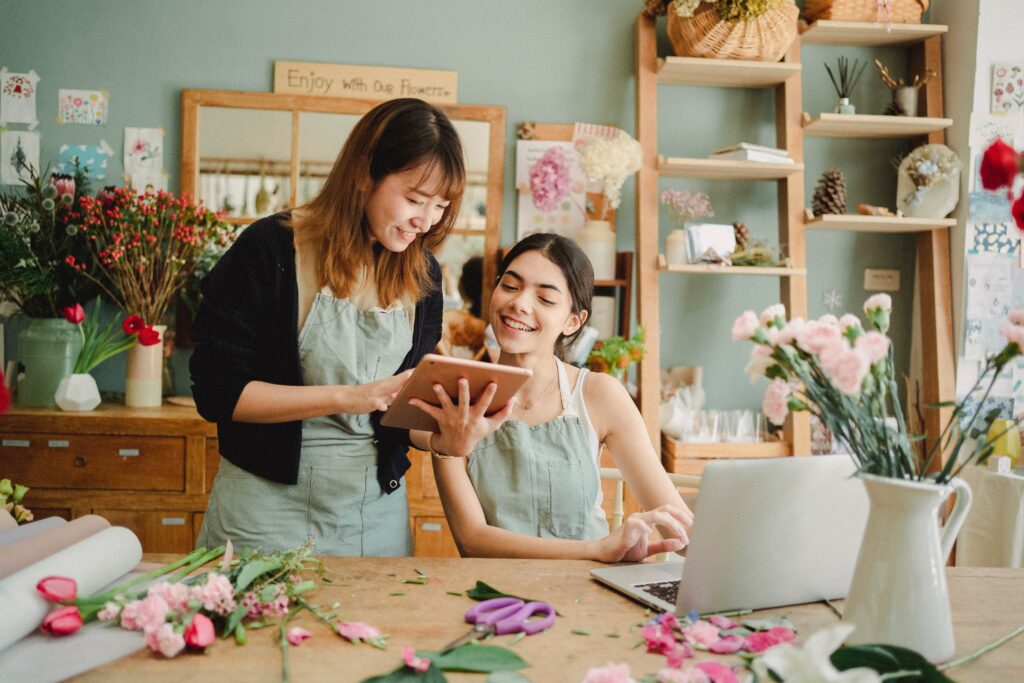Floral shop owner sitting at a table working on a computer with assistant. Lots of flowers on the table.