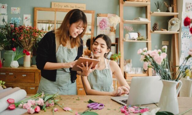 Floral shop owner sitting at a table working on a computer with assistant. Lots of flowers on the table.