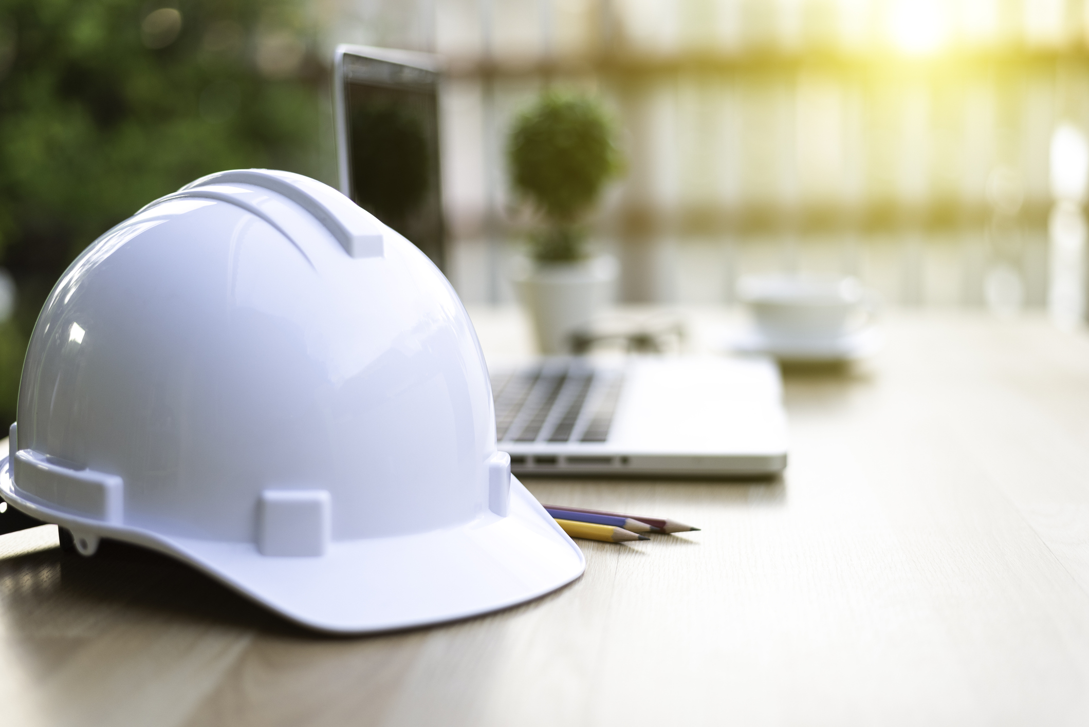 White safety helmet for Engineer sitting on a desk with a laptop and small plant in the background.