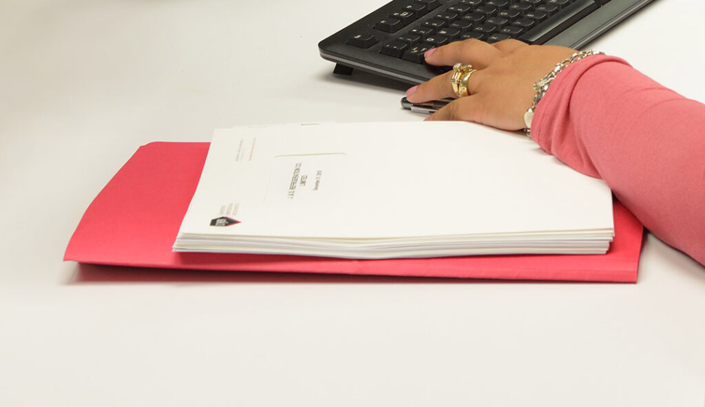 Close-up of red file folder and paper work on a white desk. Close-up of female hand wearing a red shirt using a keyboard.