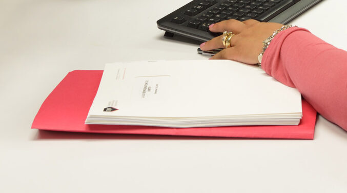 Close-up of red file folder and paper work on a white desk. Close-up of female hand wearing a red shirt using a keyboard.