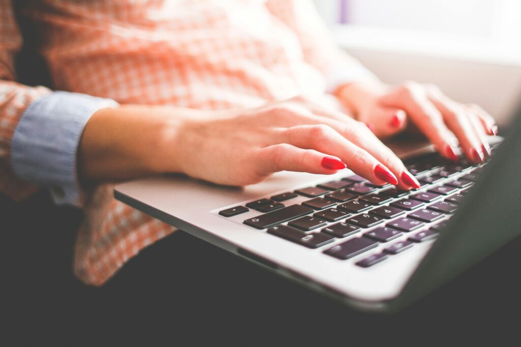 Close up of women with red nails using a laptop. The women is wearing an orange checkered shirt and jeans.