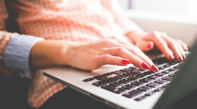 Close up of women with red nails using a laptop. The women is wearing an orange checkered shirt and jeans.