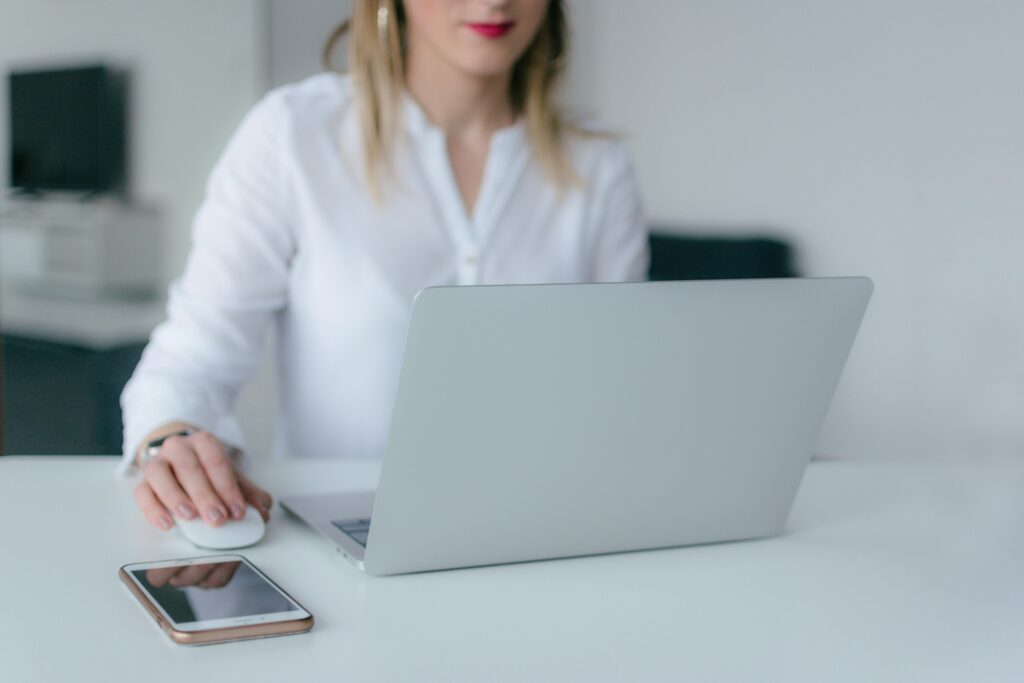 Close up of ladies hand using a mouse, sitting at a desk in front of a laptop. The table is white. The lady is wearing a white shirt.