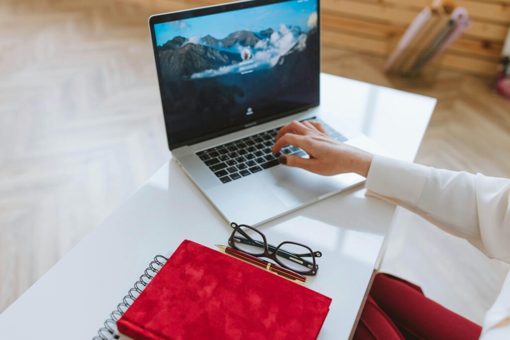 Close up of a woman's hand typing on a laptop. The woman is wearing red pants & a white shirt. A red notebook and glasses are on the table beside the laptop.